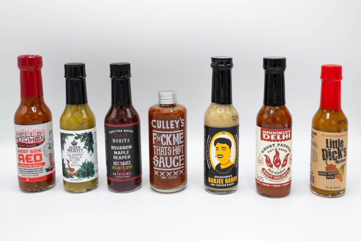 Seven hot sauce bottles of varying colors are lined up against a white background.