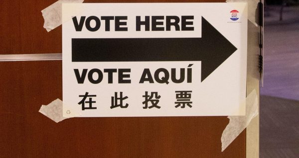 A sign that reads “VOTE HERE” and “VOTE AQUI” with a black arrow pointing towards a door.