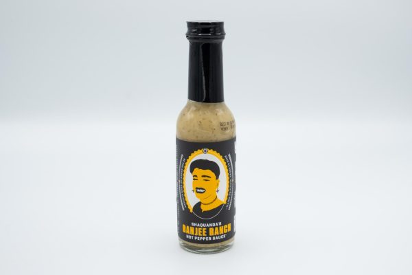 A bottle of light yellow hot sauce with a black and yellow label with a drawing of a drag queen on it, with text below that reads “Shaquanda’s Banjee Ranch Hot Pepper Sauce.”