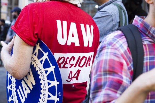 A person wearing a red shirt that reads “U.A.W. Region 9A” carries a blue circular “U.A.W.” sign.