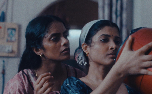 A close-up shot of two women, with one wearing a headscarf and holding an orange container, while the other is standing next to her staring at it.