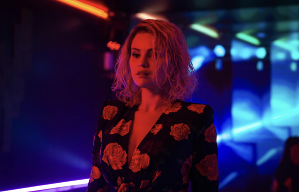 A blonde woman with bedazzled hoop earrings and a rose-patterned top stands in a room lit with neon red and blue lights.