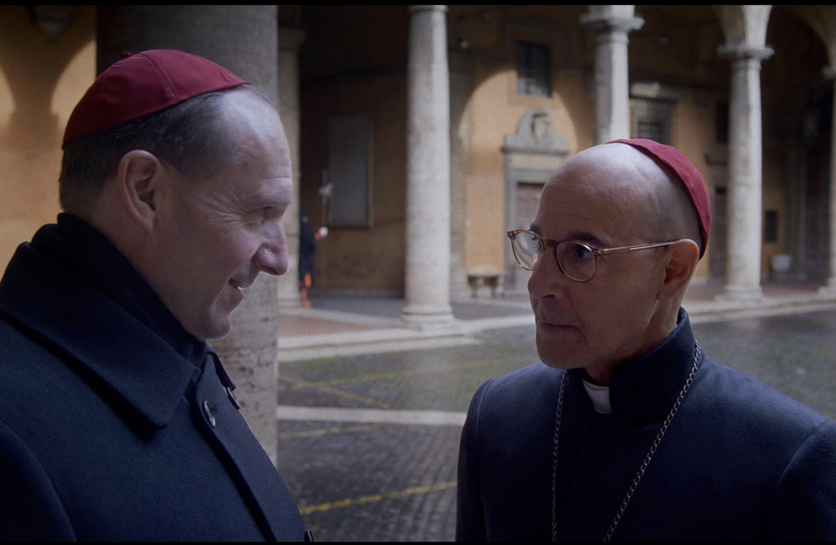 Two clergy members in red caps talking in a courtyard with stone columns and arches.