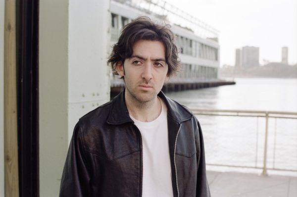 A person wearing a black leather jacket and a white shirt in front of a pier looks to the side.