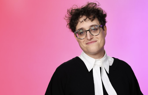 A person in a black shirt and white collar standing in front of a pink background.