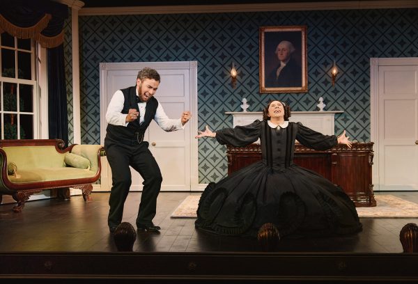 A man and woman in historical costumes on a stage set resembling an office.