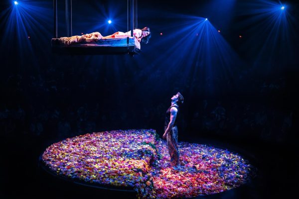 A woman hanging from a bed that is in the air, looking down at a man. Underneath them is a bed of flowers.