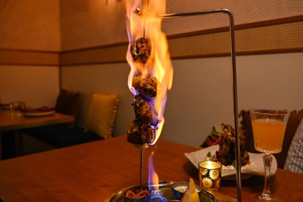 A hanging skewer of chicken is covered in orange and blue flames. It is served with lemon and other vegetables.