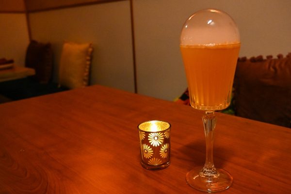 An orange cocktail with a decorative bubble of smoke on top is next to a candle on a table.