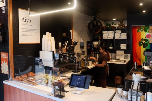 A barista makes a drink in a cafe kitchen filled with machines, ingredients and a pride flag. On the wall is a hanging poster with the word “Aiyo.”