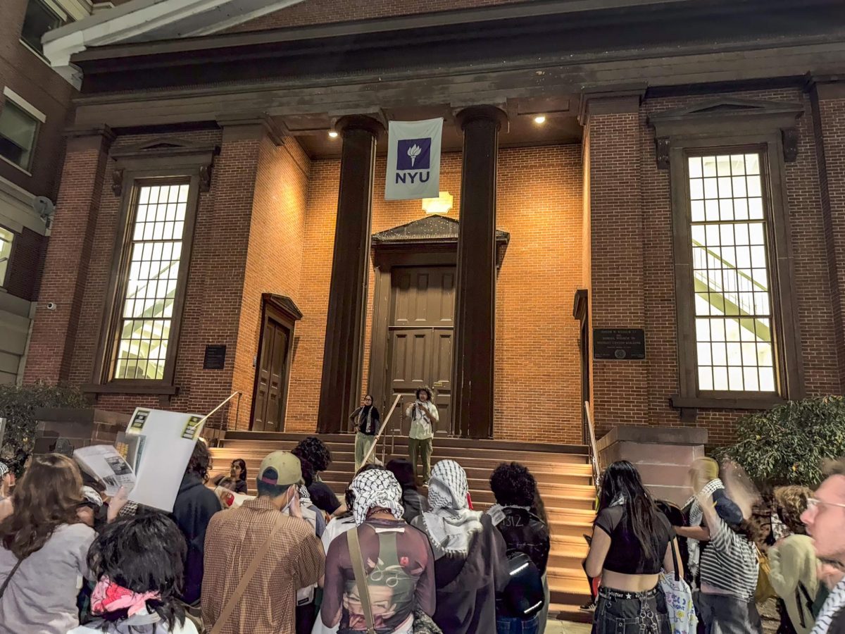 A group of people gathered in front of a building with an N.Y.U. flag. Two people stand on the stairs in front of them, one of them reading aloud from their phone.