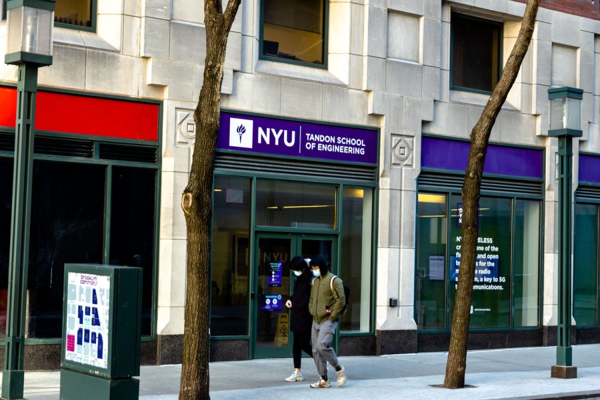 Two people walk past the entrance of an N.Y.U. building that says “TANDON SCHOOL OF ENGINEERING” on a purple background.