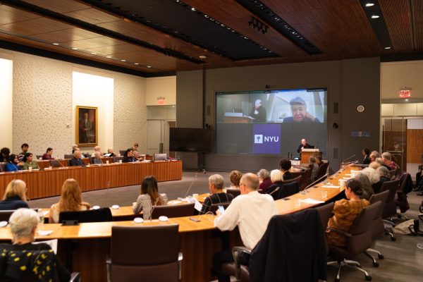 An audience sitting in rows of wooden desks attentively listening in a well-lit conference room with large windows in the background.