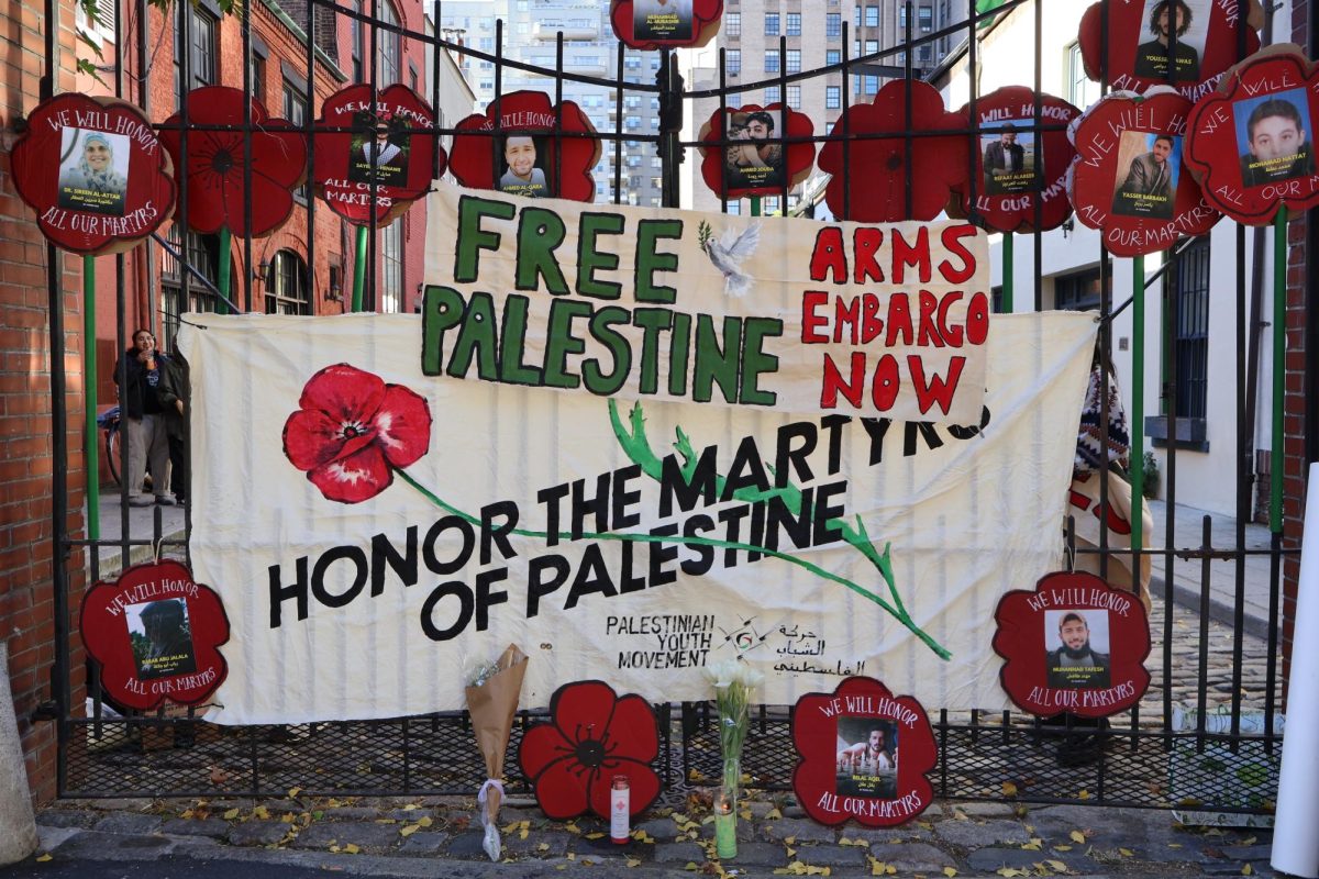 Large signs and cutouts of red flowers with photos of people in their centers hang on a fence. The largest sign says “HONOR THE MARTYRS OF PALESTINE” and a smaller sign above says “FREE PALESTINE” in green and “ARMS EMBARGO NOW” in red.