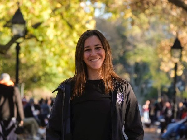 A woman stands in a park wearing a black jacket with a purple N.Y.U. logo and smiles at the camera.