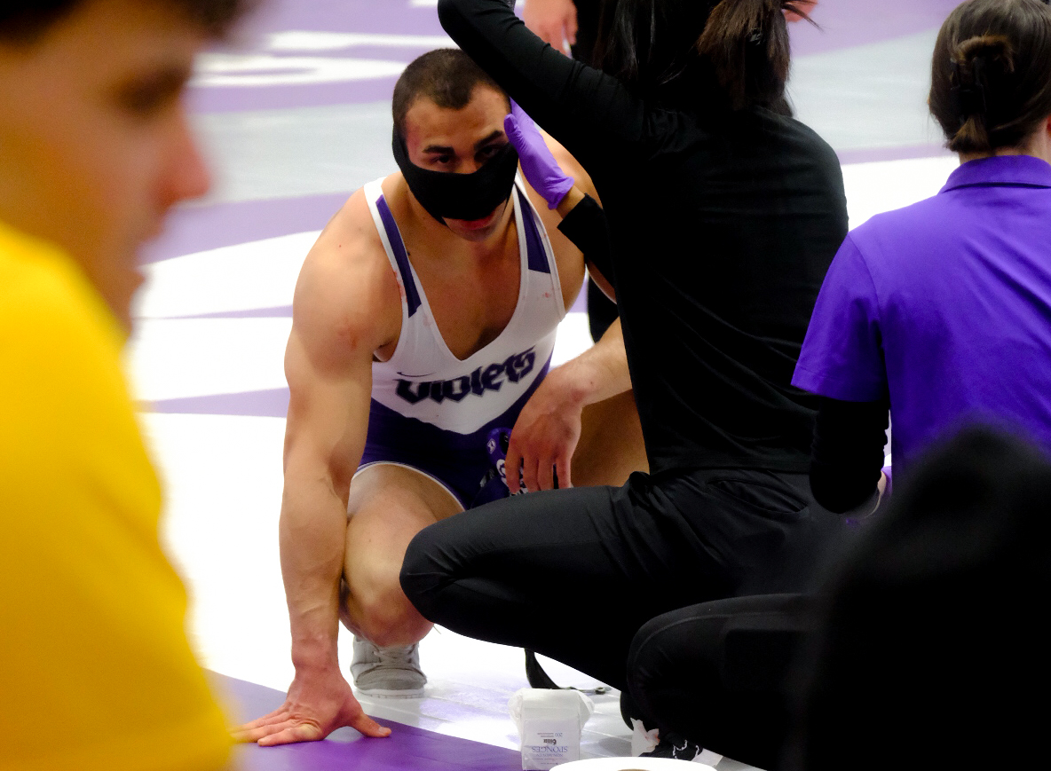 A medic applies a black bandage to an N.Y.U. wrestler crouched on the ground in a purple leotard that reads “VIOLETS.”