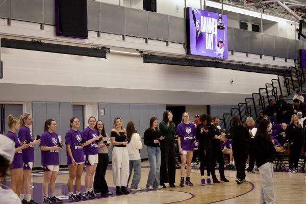 A group of people line up in the middle of a basketball court, many of whom wear a purple and white N.Y.U. T-shirt, basketball shorts and Nike sneakers.