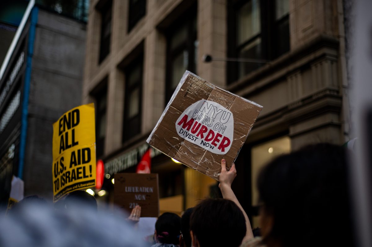 A person holding a brown sign that says, “NYU RUNS ON MURDER DIVEST NOW.”