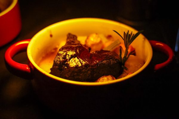 A beef short rib on top of root vegetables in a bowl with red handles.