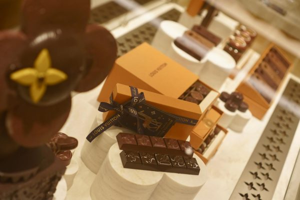 A shot of various orange boxes with a ribbon tied around one of them. The words “LOUIS VUITTON” are printed on the ribbon. Displayed next to the boxes are various chocolate squares and truffles.