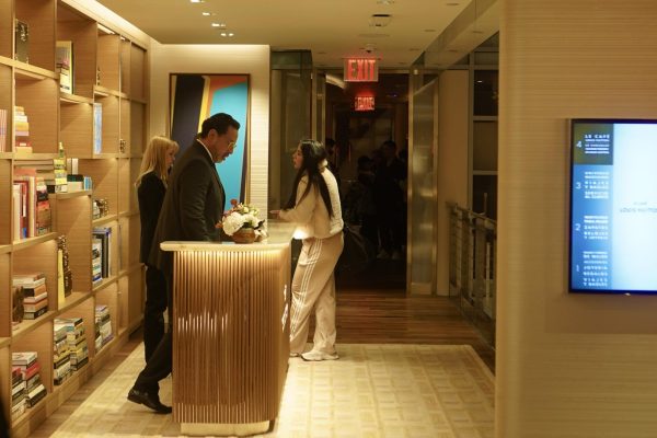 A golden room with a floor-to-ceiling bookshelf behind a reception desk. Receptionists in formal clothing stand behind the desk, while a woman in street clothes stands in front.