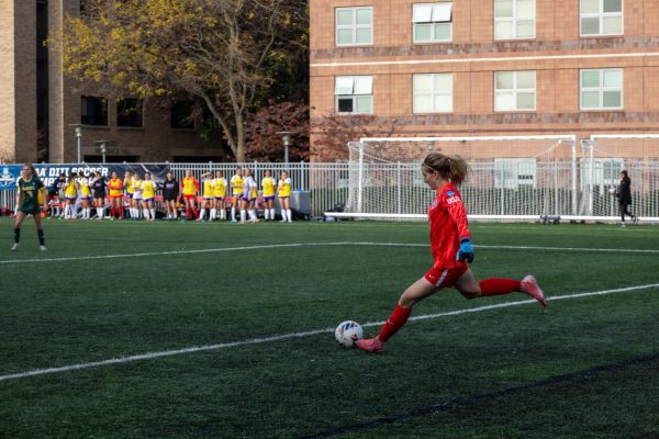 A student athlete goalkeeper in a red uniform prepares to kick a soccer ball.