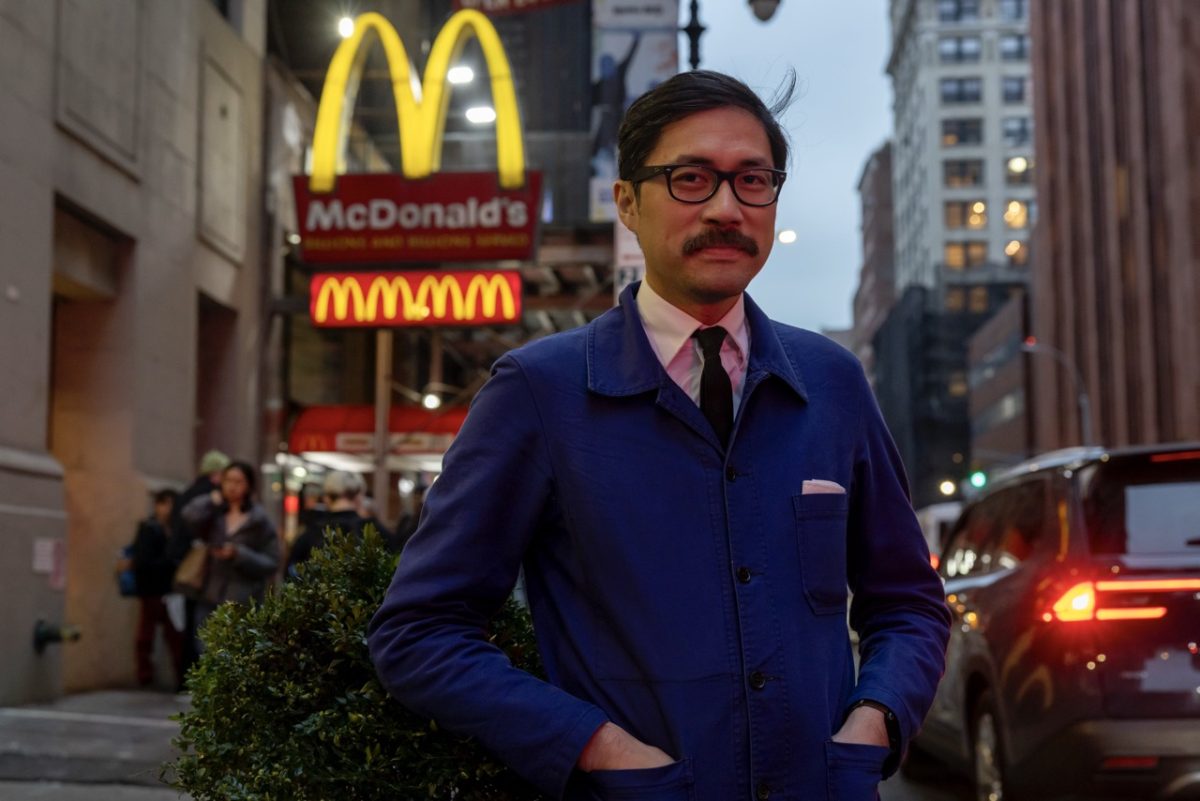 A man with glasses and a mustache wearing a blue jacket stands in front of the McDonald’s golden arches.