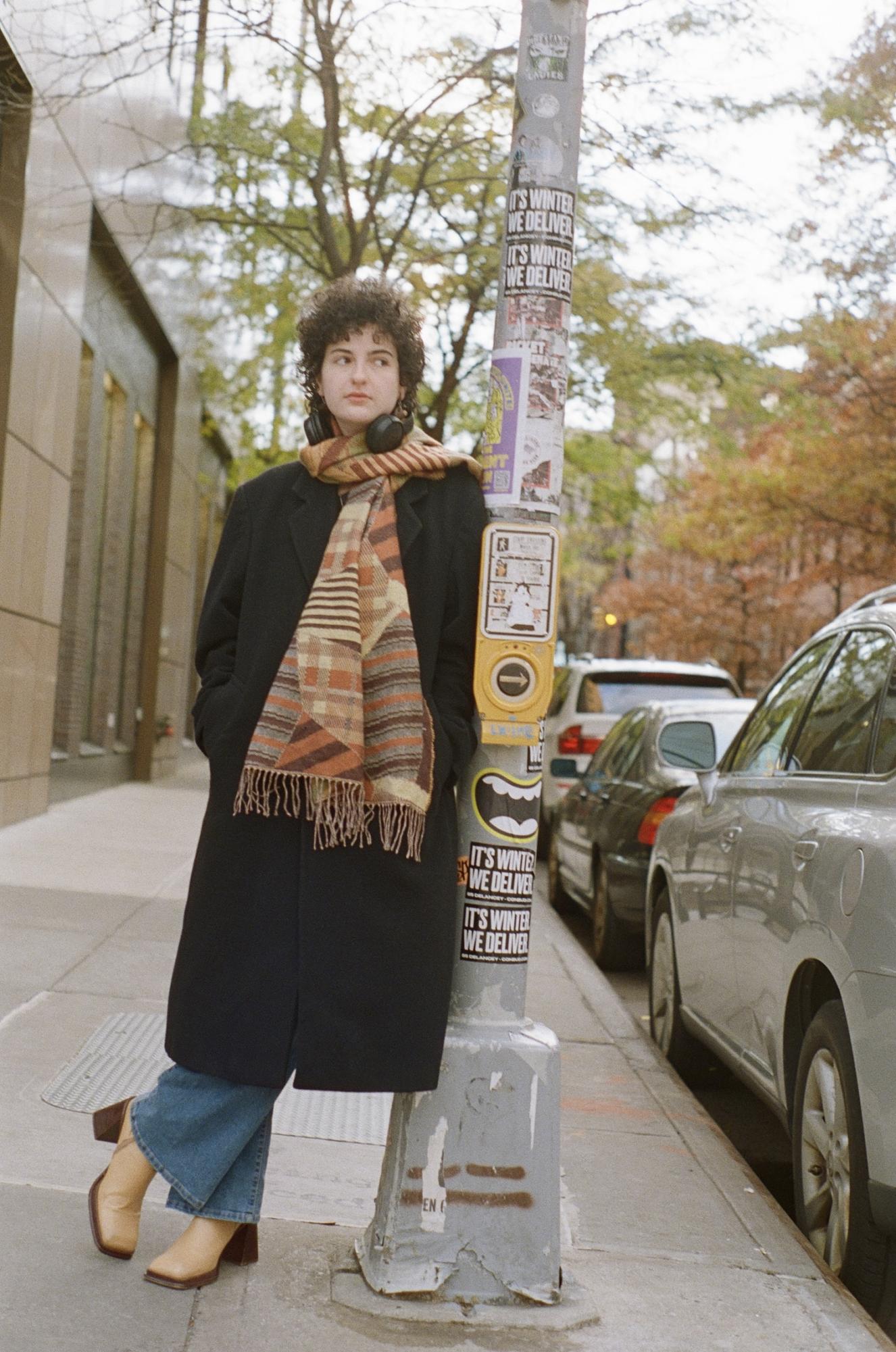 A person in a large dark coat and an orange scarf leans against a pole.