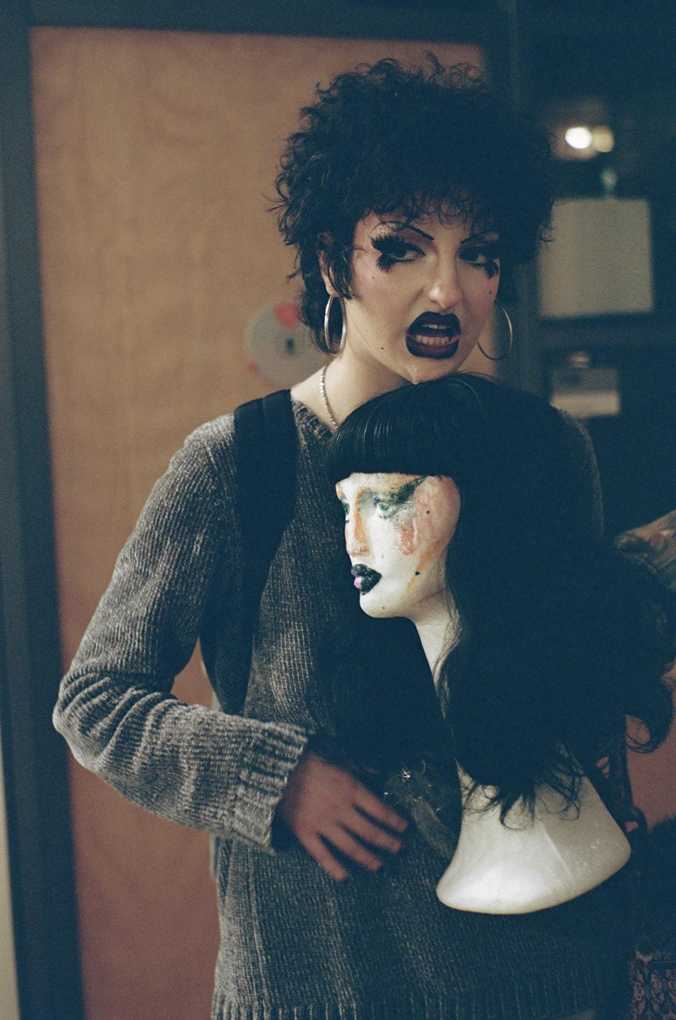 A person in elaborate makeup wearing a sweater holds a mannequin head with a wig.