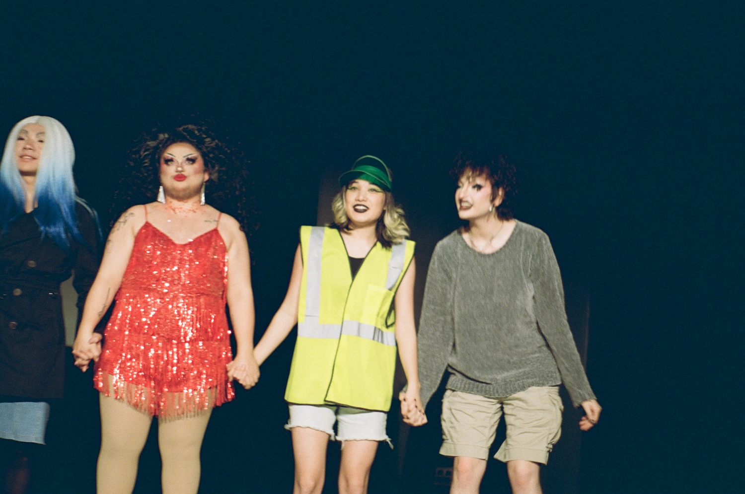 Four people in costumes and makeup stand onstage, holding hands.