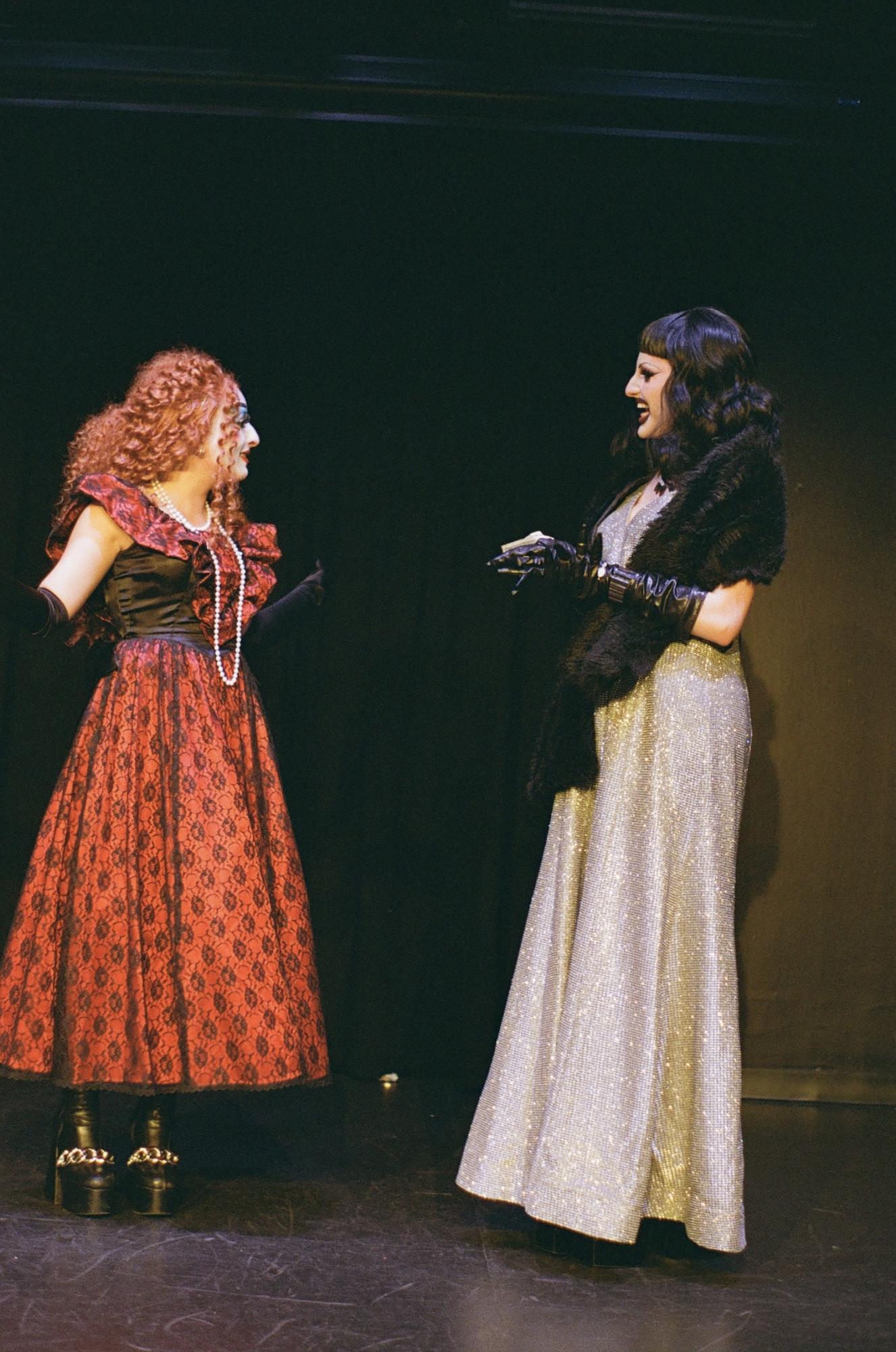 Two people in costumes and dramatic makeup stand on a stage facing each other. The person on the left wears a red and black patterned dress; the person on the right wears a black fur shawl and long silver dress.