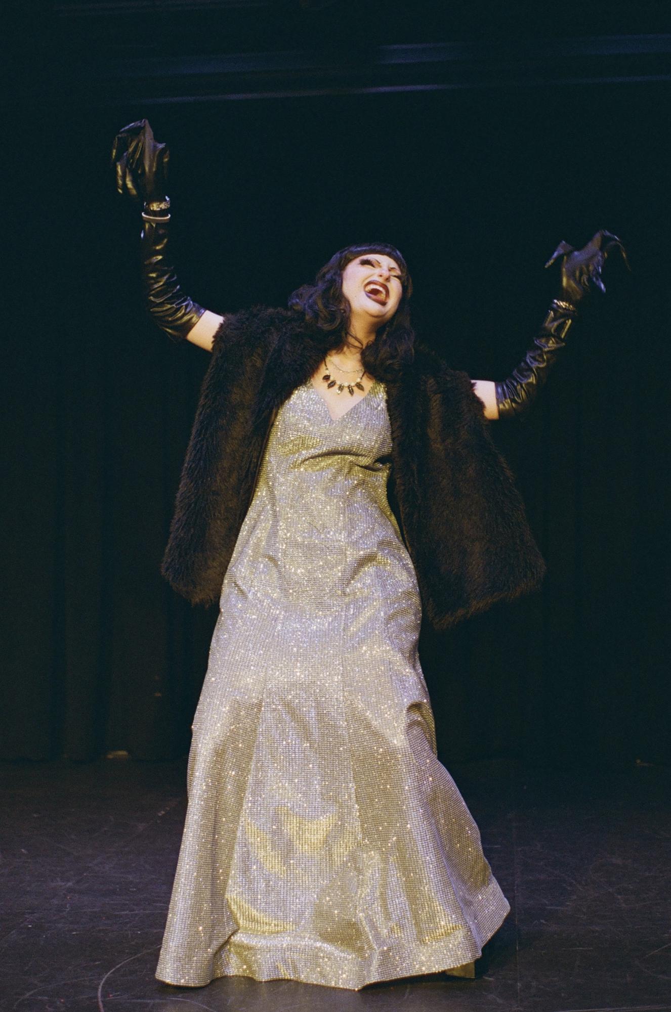 A person wearing a long silver dress, a black fur shawl and black gloves stands onstage, singing with their head thrown back and arms up.