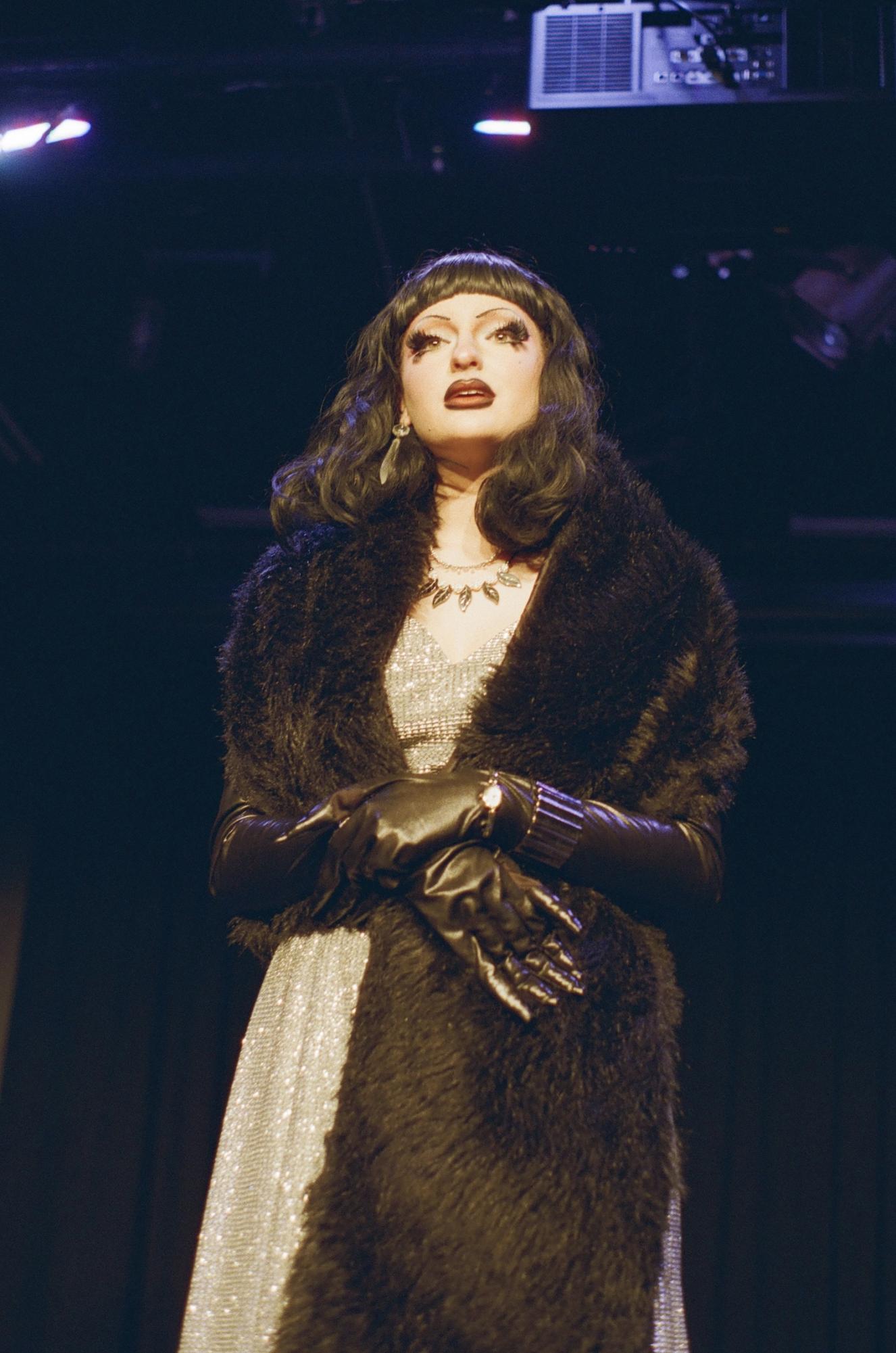 A person stands on stage staring into the distance. They have dark hair and wear a black fur shawl, black gloves and a silver dress.