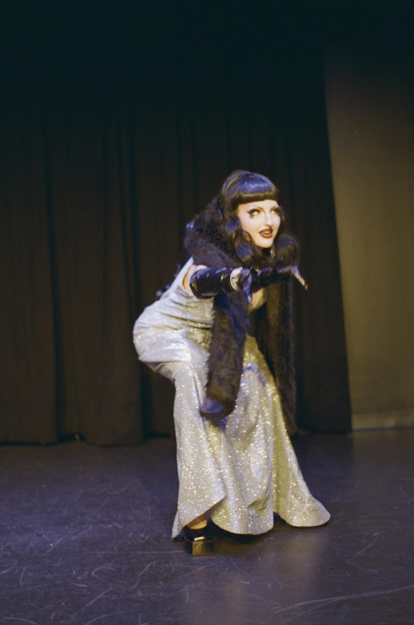 A person wearing a long silver dress and a black fur shawl stands onstage, pointing out towards the audience.