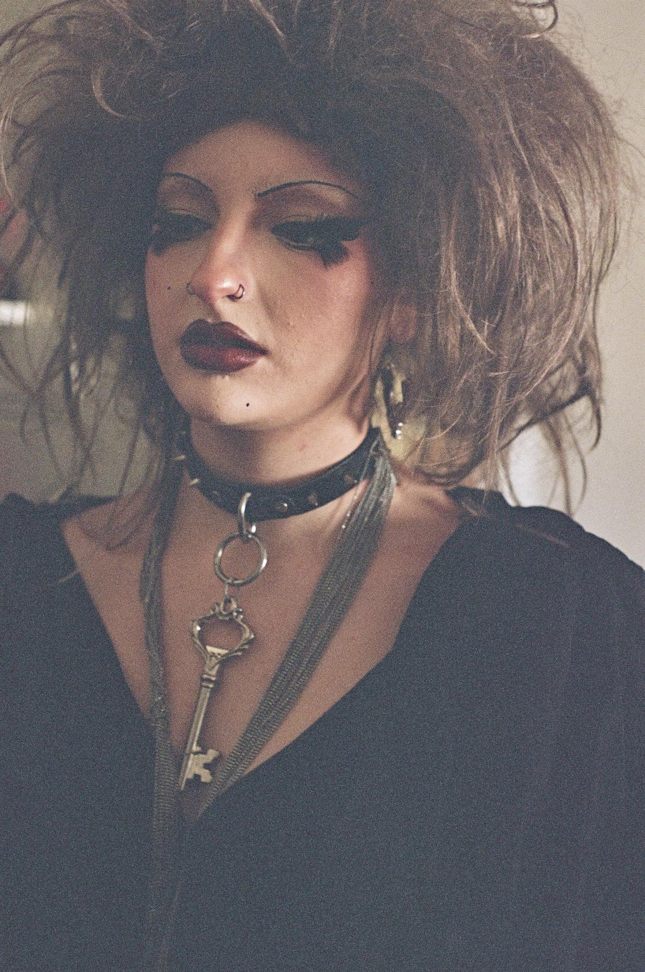 A person with dramatic makeup and wild brown hair wearing a studded choker necklace with a key on it and a thin silver scarf.