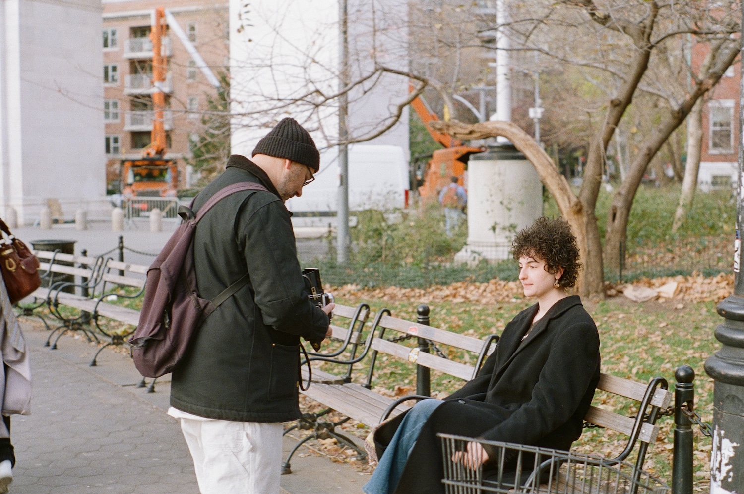 An older man takes a picture of a person in a dark coat sitting on a park bench.