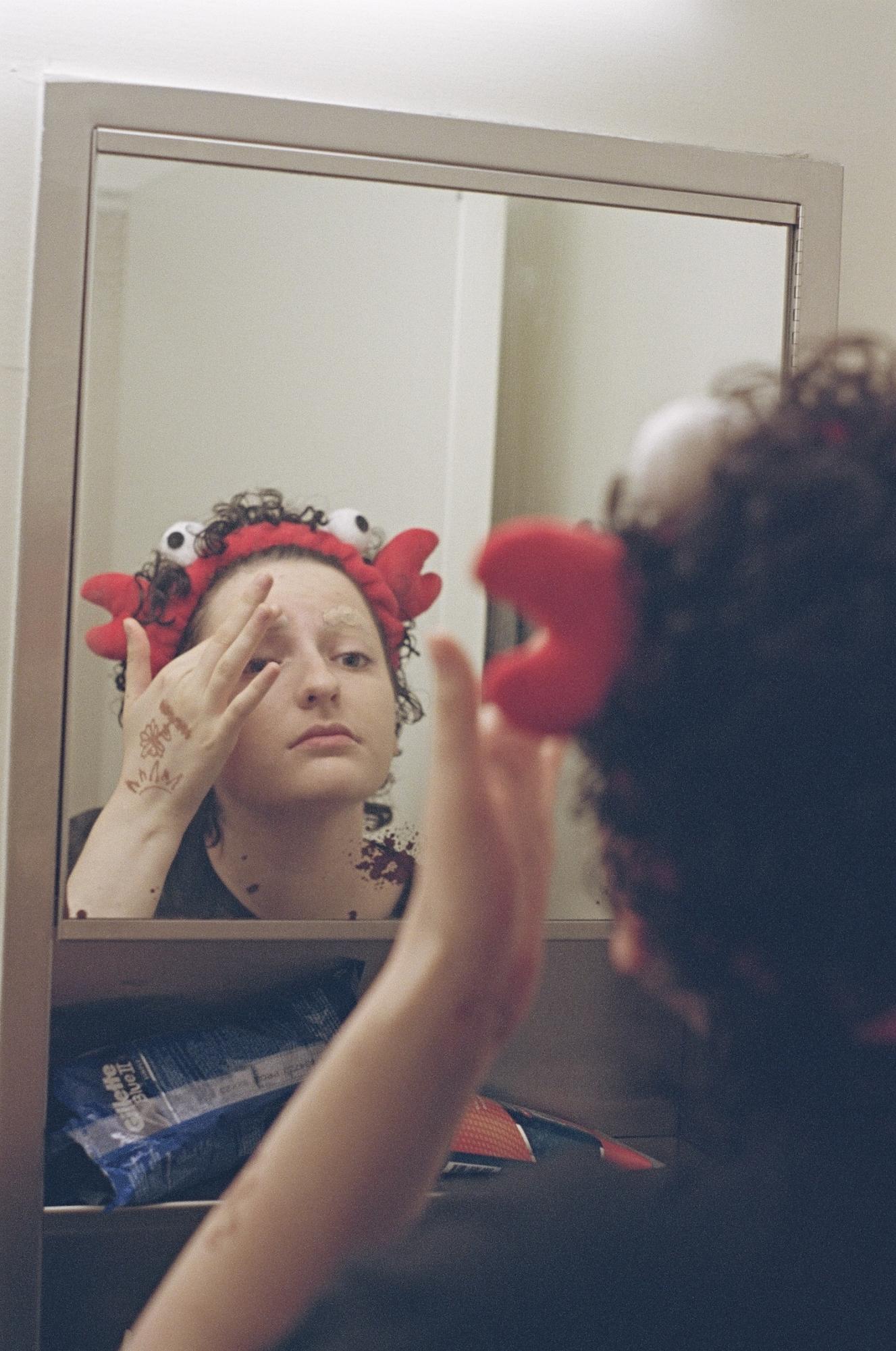 A person wearing a red crab headband does their eyebrow makeup in a mirror.