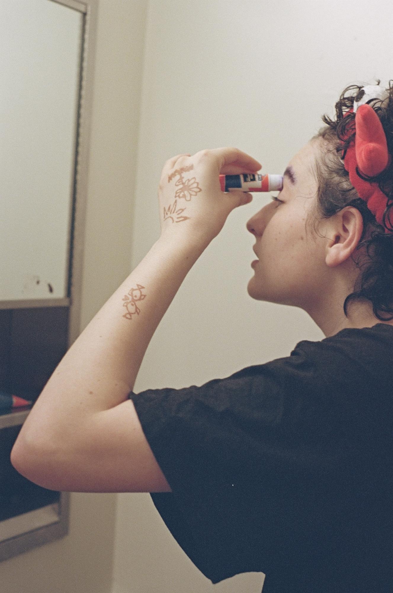 A person wearing a black T-shirt and a red headband stands in front of a mirror, putting glue on their eyebrows.