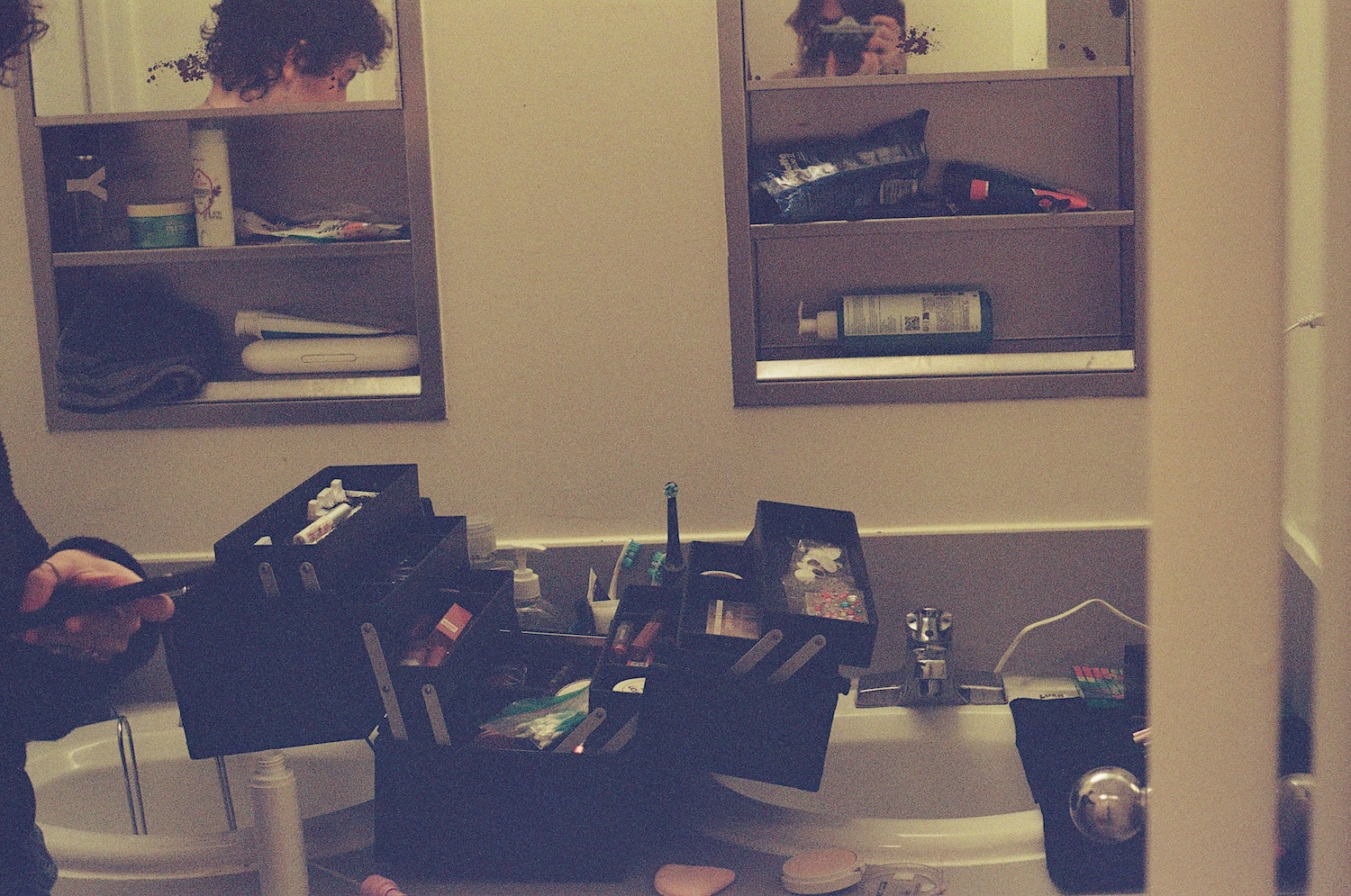 A cluttered bathroom counter with an assortment of makeup products in a large black box. In the mirrors, a person's face and another person holding a camera are visible