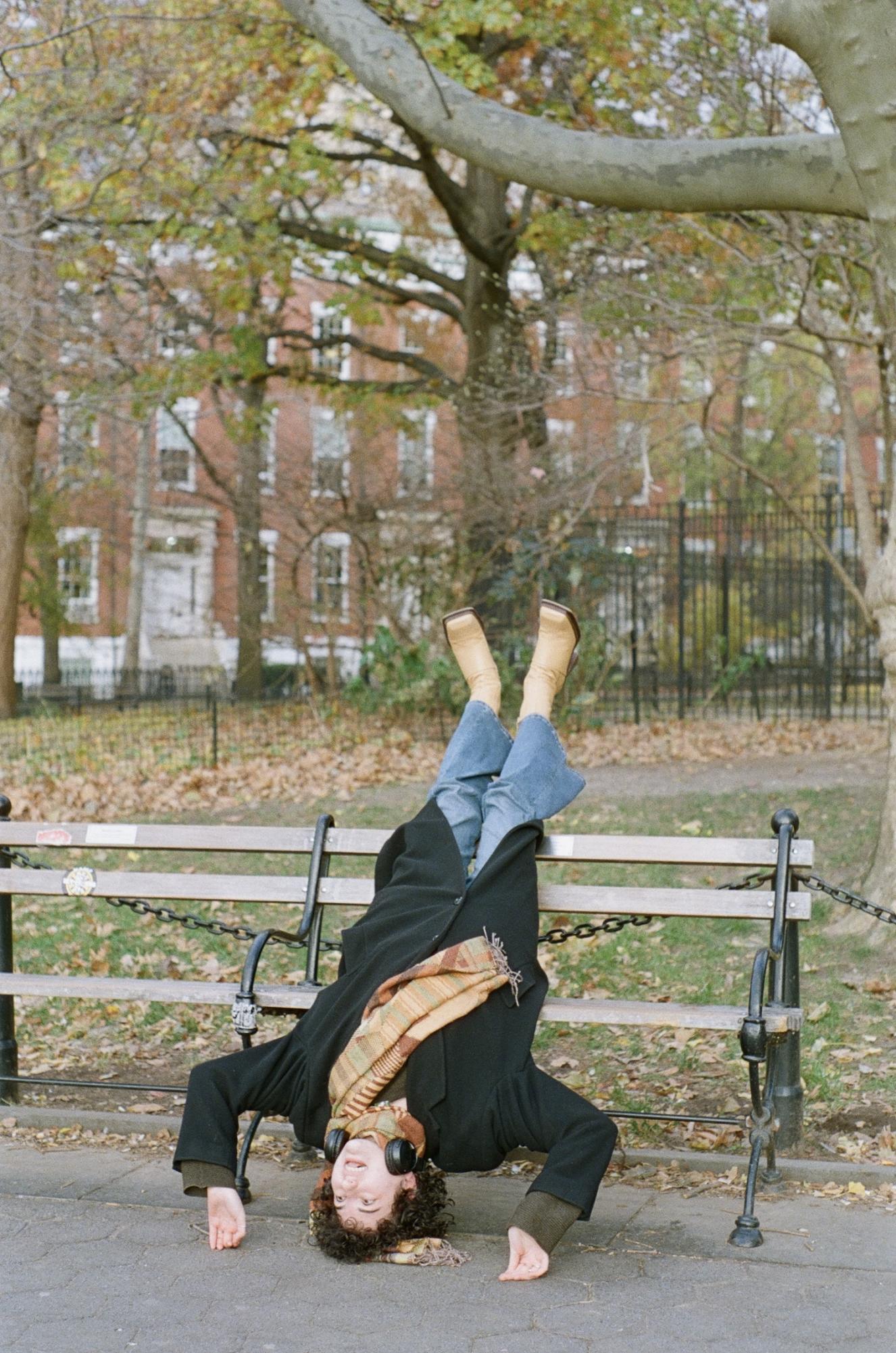 A person in a dark coat and an orange scarf with headphones around their neck lays upside down on a park bench, their head on the ground and their feet in the air.