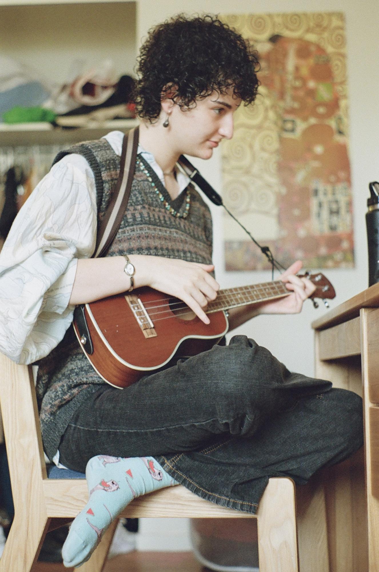 A person wearing a sweater vest sits in a chair, playing the ukulele.