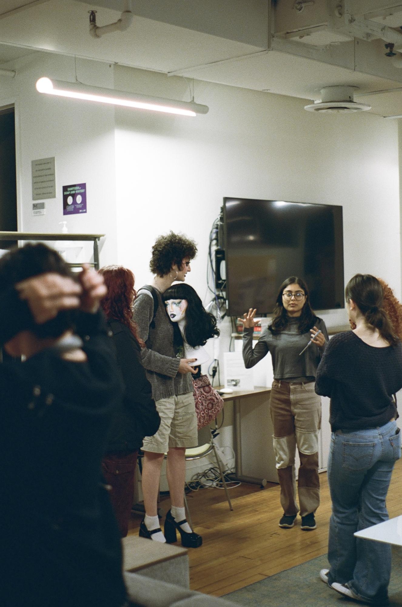 A person talking to a group of four people inside a small room. Next to them stands another person holding a mannequin head with a wig.