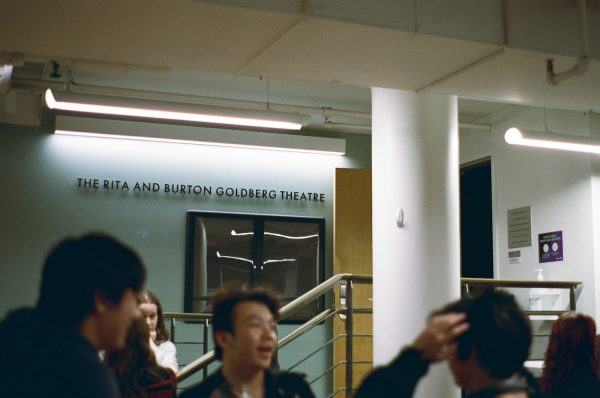 On a wall above a crowd of people is a sign saying “THE RITA AND BURTON GOLDBERG THEATRE.”