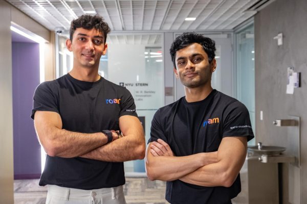 Two men in black T-shirts with an orange and blue “roam” logo printed on it stand in a hallway, smiling with their arms crossed.