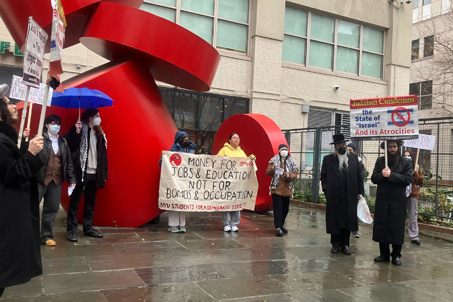 A group of protesters hold signs in front of a large red sculpture. Some signs read “Judaism Condemns the State of ‘Israel’ And its Atrocities” and “MONEY FOR JOBS AND EDUCATION NOT FOR BOMBS AND OCCUPATION” above “N.Y.U. STUDENTS FOR A DEMOCRATIC SOCIETY.”