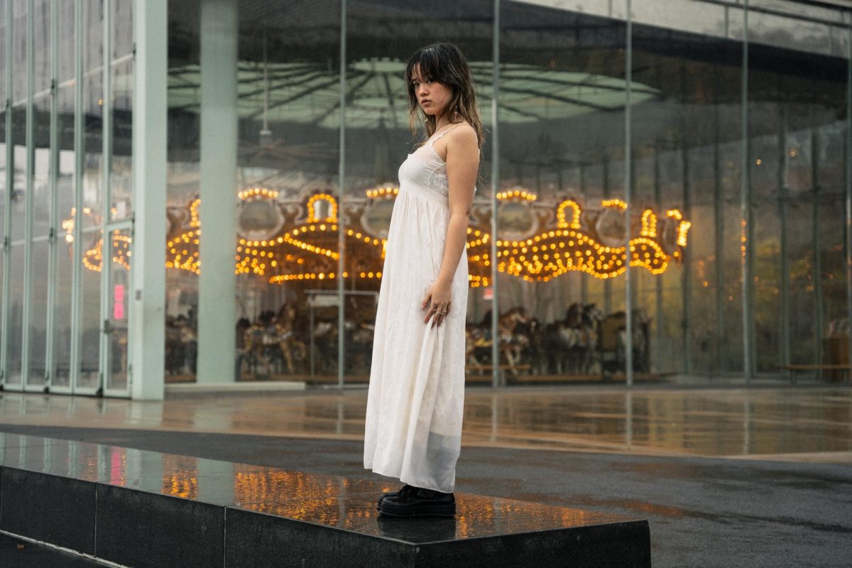 A woman standing in a long, white dress in front of a building with a carousel behind her.