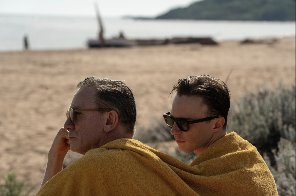 Two men sit next to each other on a beach, a yellow blanket wrapped around their shoulders.