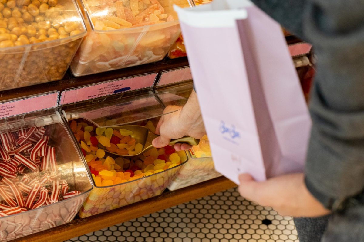 A hand scooping “Tutti Frutti Original” candy from a large container. Around it are rows of containers of candy.