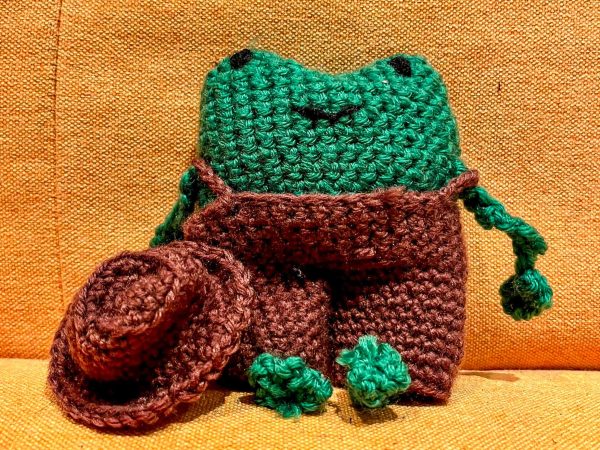 A green crocheted frog wearing brown overalls sitting on a light brown surface. Next to it is a crocheted brown hat.