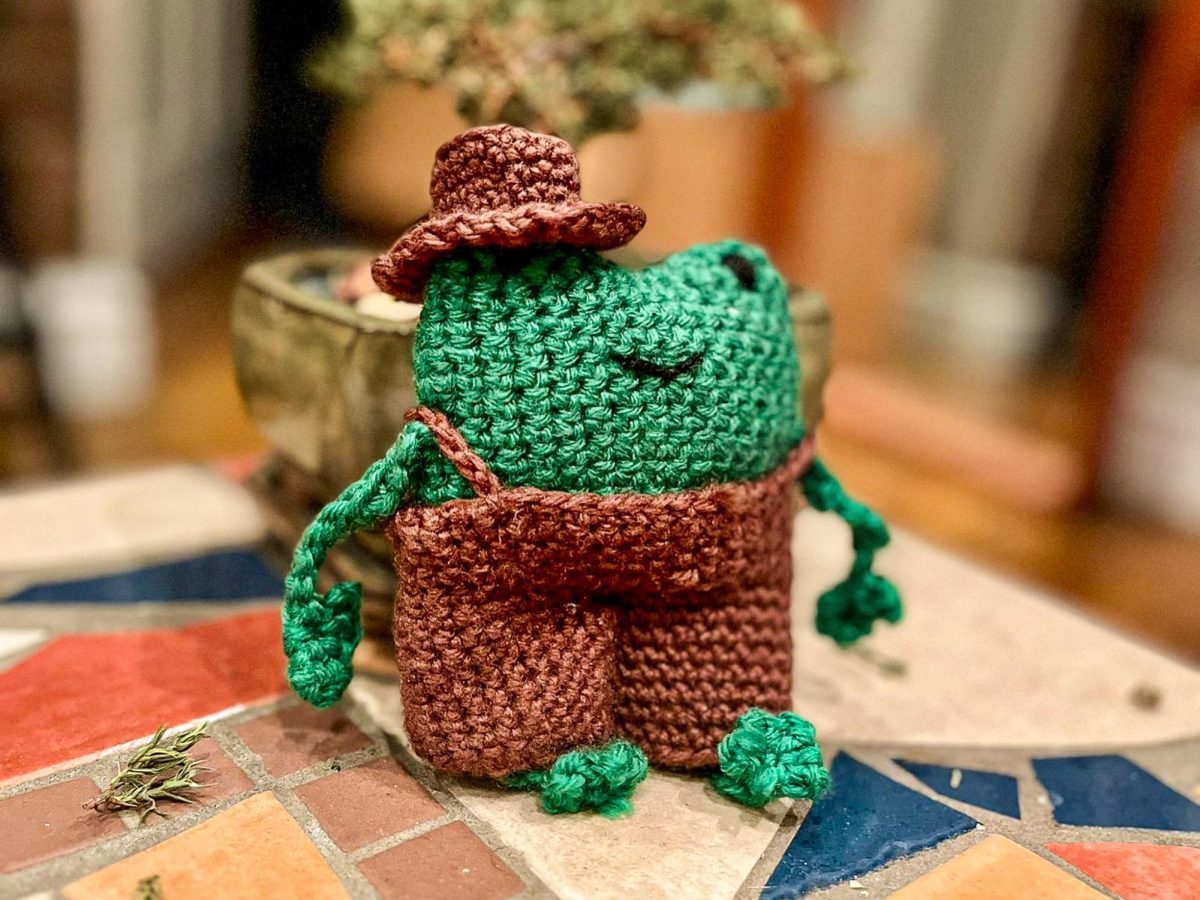 A green crocheted frog wearing brown overalls and a small brown hat sitting on a tiled surface.